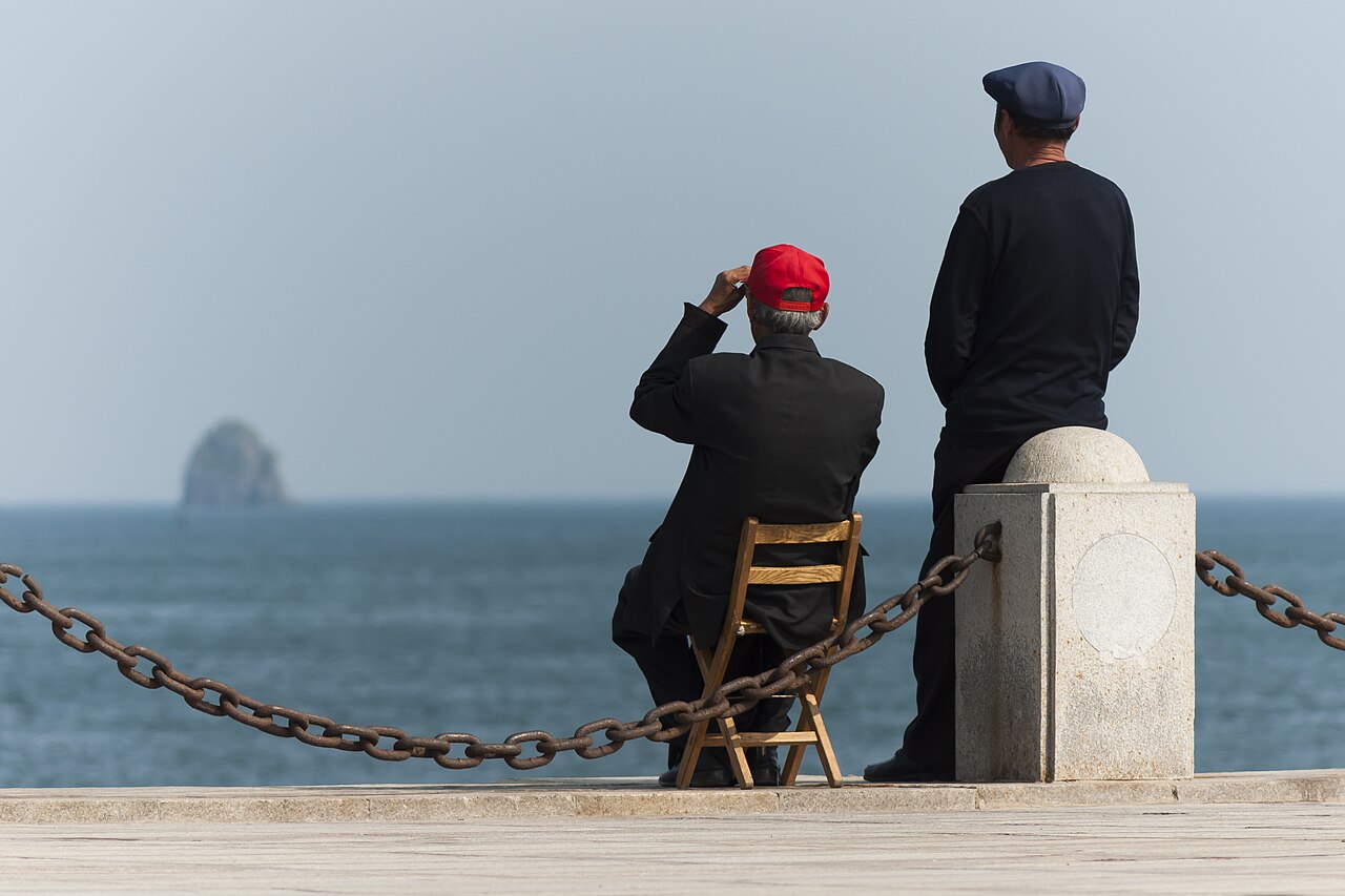 Des habitants sur la promenade maritime de la baie de Xinghai, avec un petit îlot émergeant des eaux calmes derrière eux.