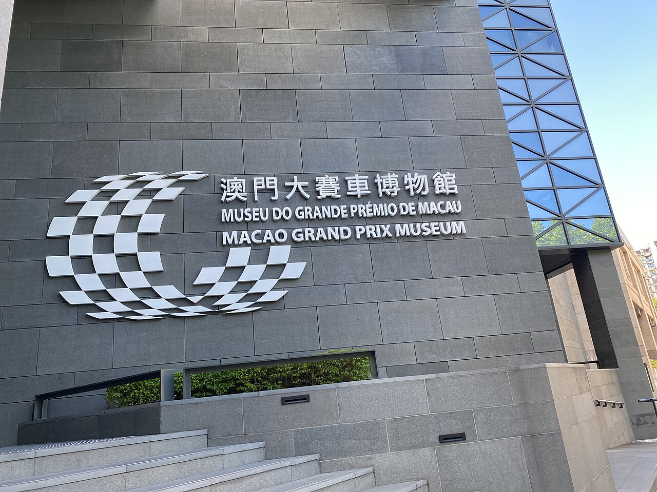 Die graue Steinfassade des Macao Grand Prix Museums mit einem Schachbrettfahnen-Emblem und dem Namen des Museums auf Chinesisch, Portugiesisch und Englisch.
