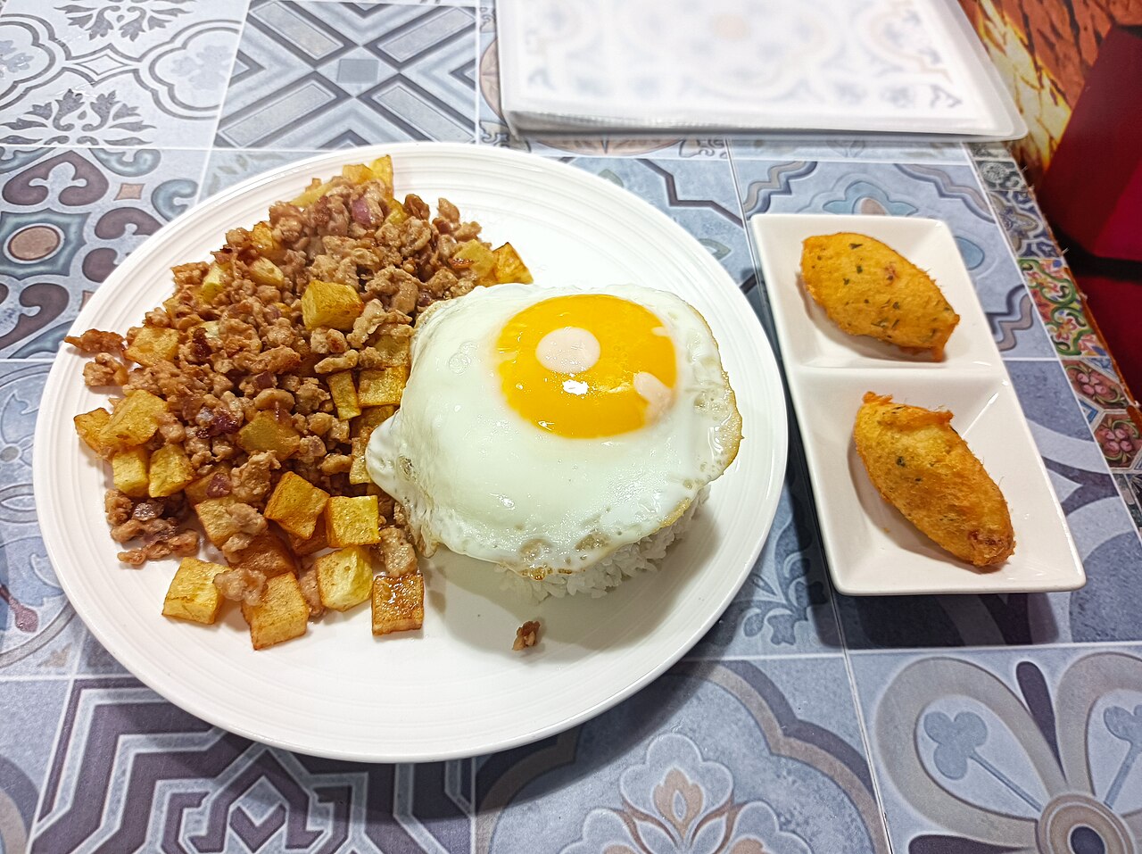 Schweinehack und gebratene Kartoffelwürfel neben einem Spiegelei auf Reis, mit zwei goldenen Kroketten als Beilagen auf einem gemusterten Kacheltisch.