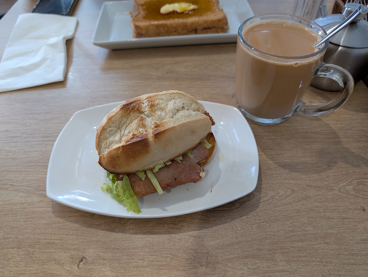 Ein Schweinekotelett in einem knusprigen, aufgeschnittenen Brötchen auf einem weißen Teller, dahinter ein Glas Milchtee und eine Scheibe Honigtoast.