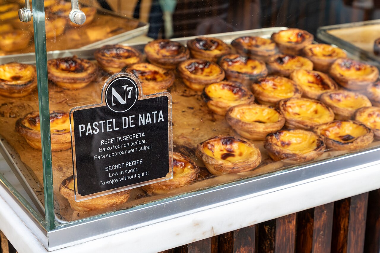 Tabletts mit Eiertörtchen hinter der Bäckereischeibe, ihre Puddingoberflächen blasig und karamellisiert, neben einer Karte mit der Aufschrift Pastel de Nata.