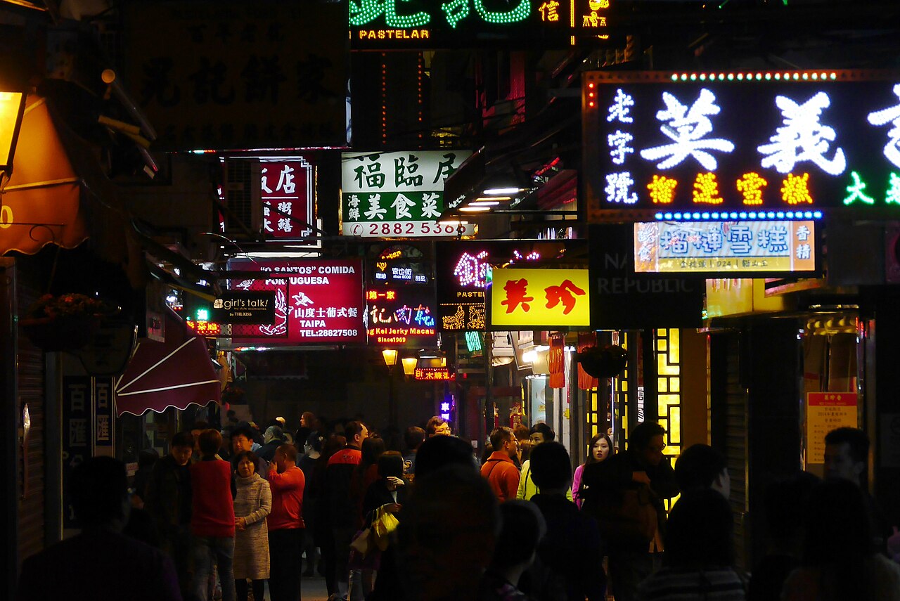 A narrow lane packed with people under walls of neon and lightbox signs in Chinese and Portuguese, bakery and jerky shops on both sides.