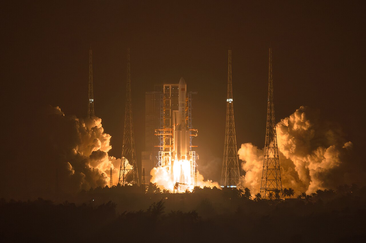 Un cohete Larga Marcha 5 despegando de noche desde Wenchang, con la rampa iluminada en naranja por los gases de escape, cuatro torres pararrayos y una hilera de palmeras a su alrededor.