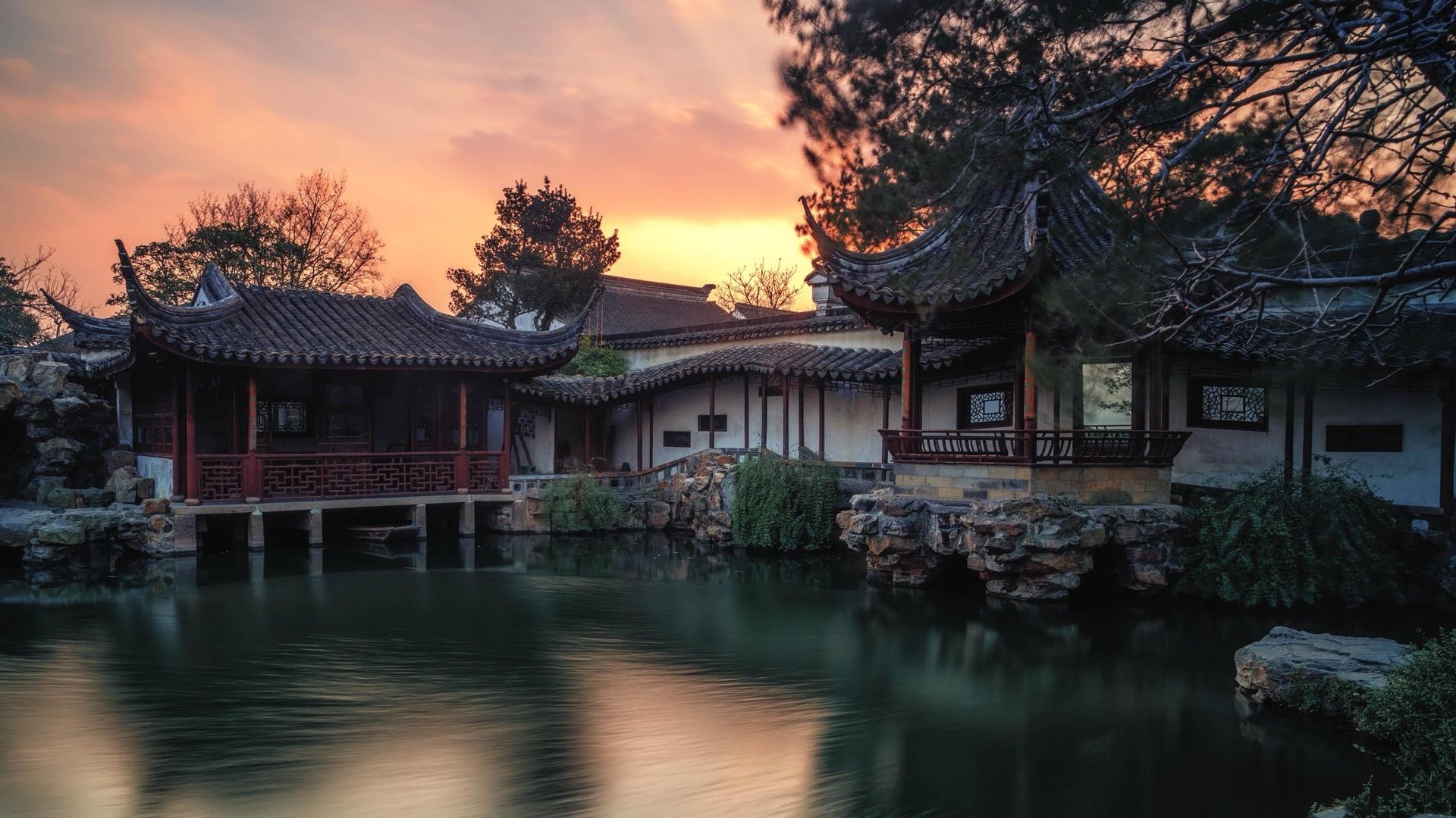A classical Suzhou garden at sunset: timber halls and an upswept pavilion roof mirrored in still water