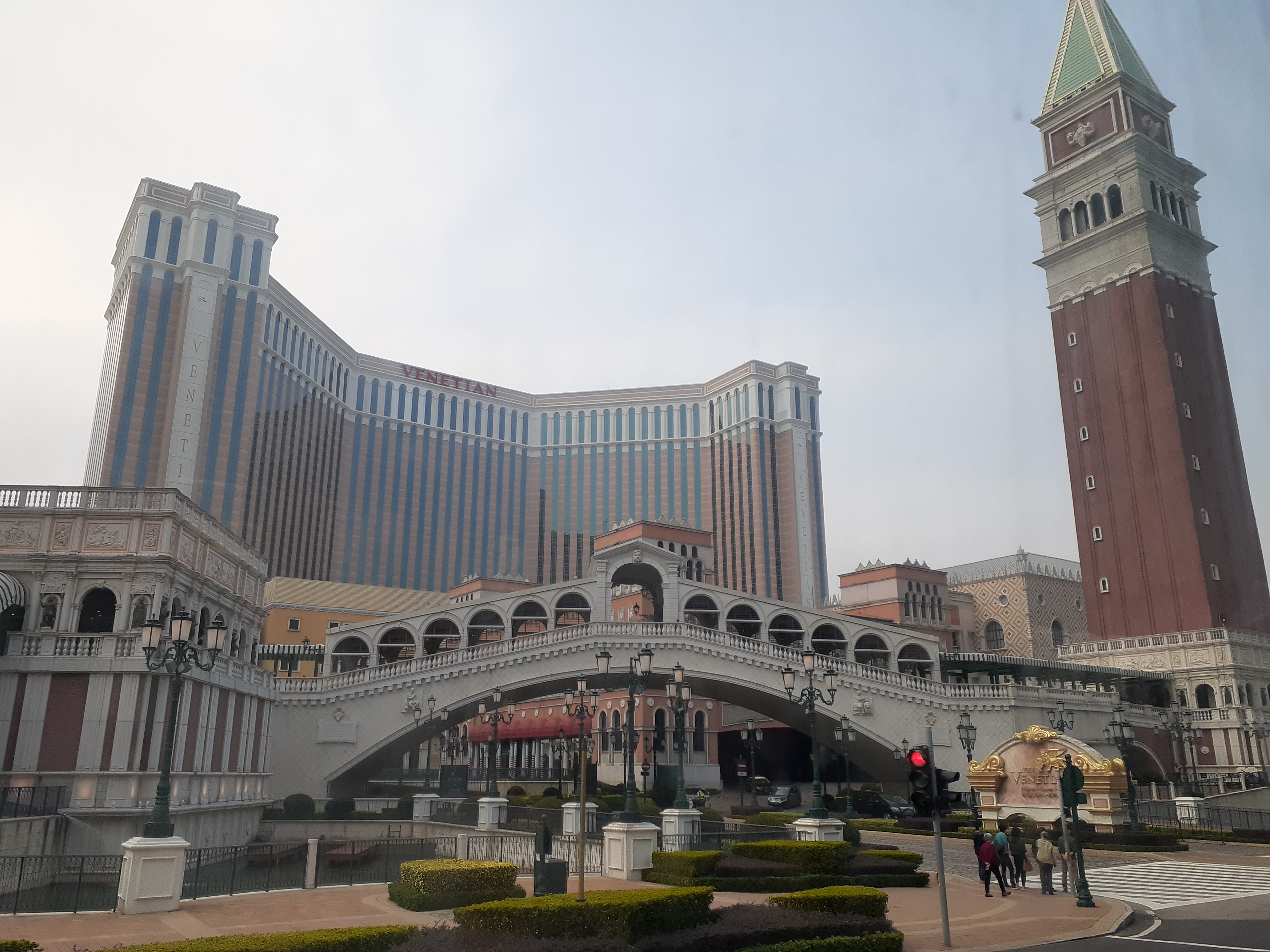 Eine Nachbildung der Rialtobrücke und ein gemauerter Glockenturm vor dem gestreiften Hotelblock des Venetian, mit geschnittenen Hecken und einem Fußgängerüberweg davor.