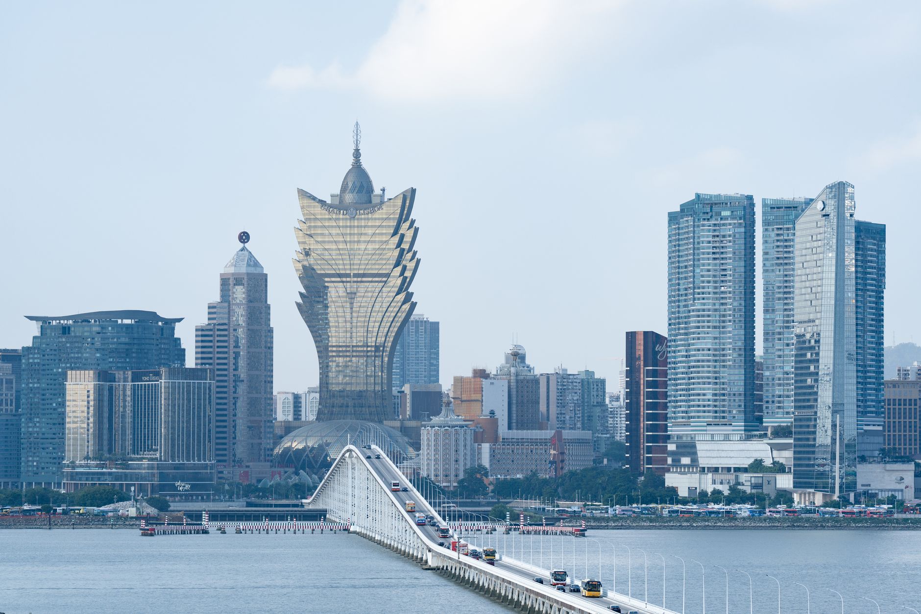 Macaus Skyline jenseits des Wassers, das goldene, lotusförmige Grand Lisboa in der Mitte, davor Busse, die die Dammbrücke überqueren.