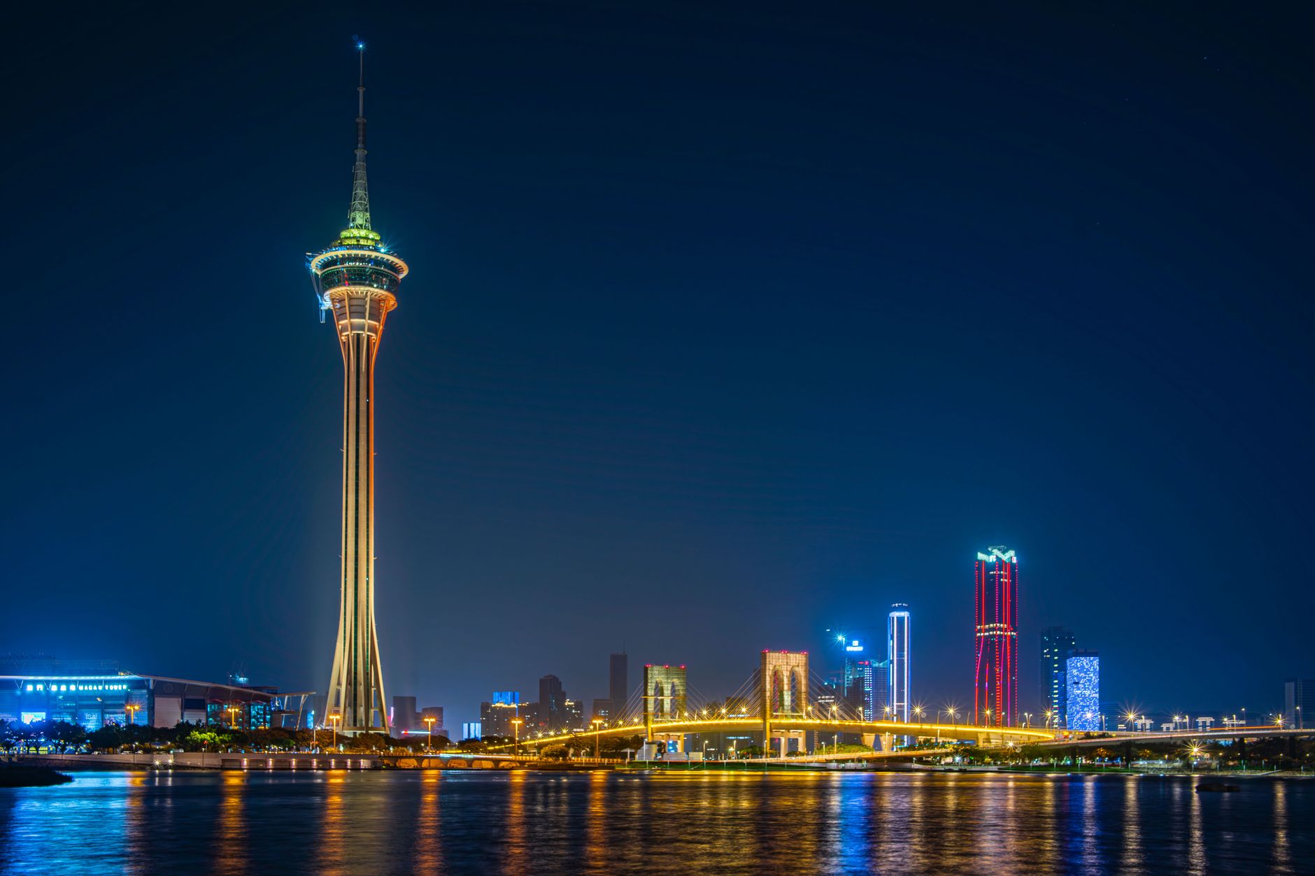 Der beleuchtete Macau Tower und sein Aussichtsring über einer Schrägseilbrücke, mit der Skyline von Cotai und ihren Spiegelungen dahinter.